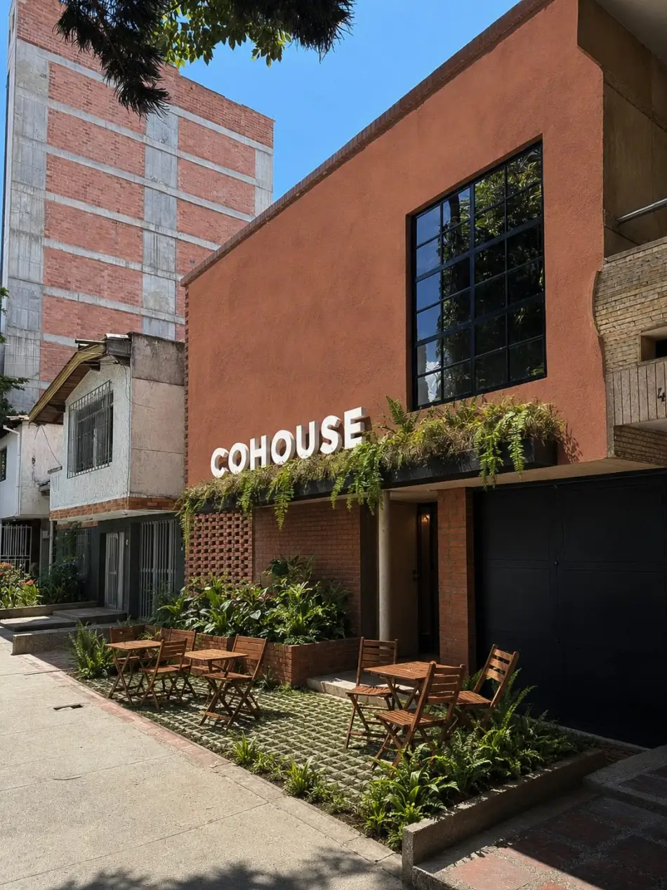 Fachada terracota de COHOUSE con letras blancas, helechos colgantes, celosía de ladrillo y mesas de café de madera sobre la acera.
